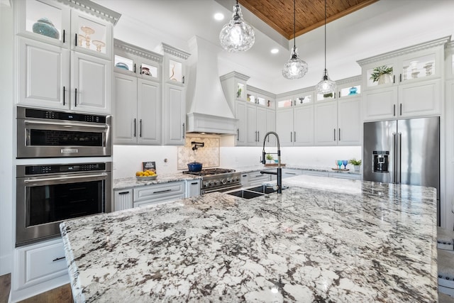 kitchen with custom exhaust hood, sink, premium appliances, white cabinetry, and light stone countertops