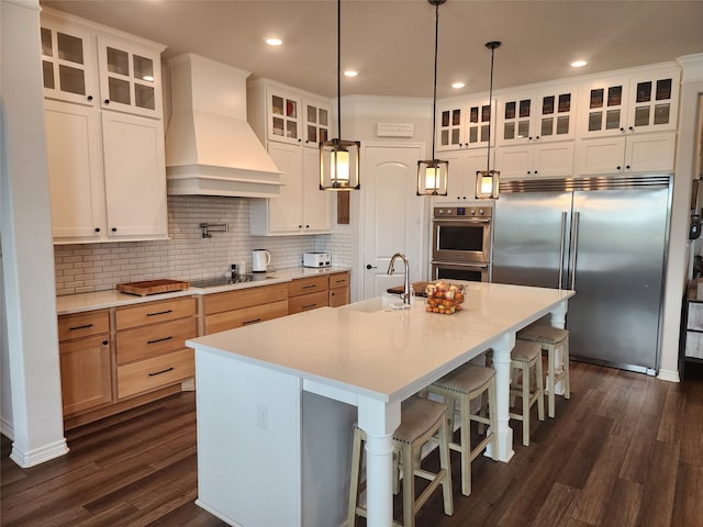 kitchen with white cabinets, hanging light fixtures, a center island with sink, stainless steel appliances, and custom range hood