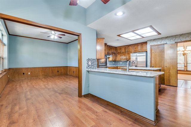 kitchen with kitchen peninsula, stainless steel fridge, light stone counters, ceiling fan with notable chandelier, and light hardwood / wood-style flooring