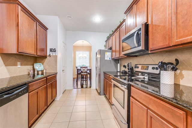 kitchen featuring decorative backsplash, dark stone countertops, a notable chandelier, light tile patterned flooring, and stainless steel appliances