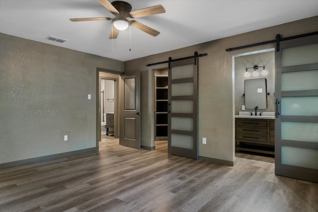 unfurnished bedroom with a barn door, ceiling fan, ensuite bathroom, hardwood / wood-style floors, and sink