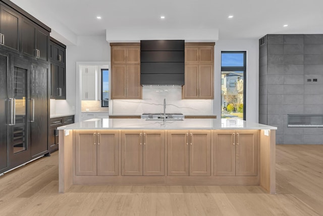 kitchen featuring a center island with sink, decorative backsplash, light wood-type flooring, built in fridge, and custom range hood