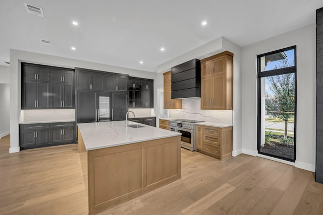 kitchen featuring high end stainless steel range oven, premium range hood, an island with sink, backsplash, and light stone countertops