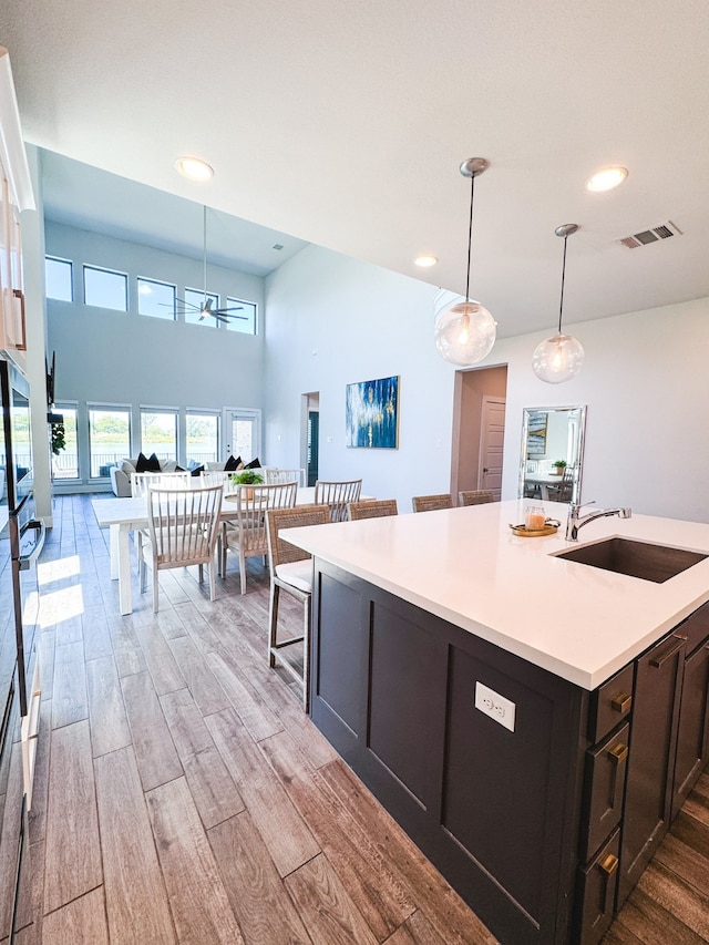 kitchen with a towering ceiling, sink, decorative light fixtures, light hardwood / wood-style flooring, and a kitchen island with sink