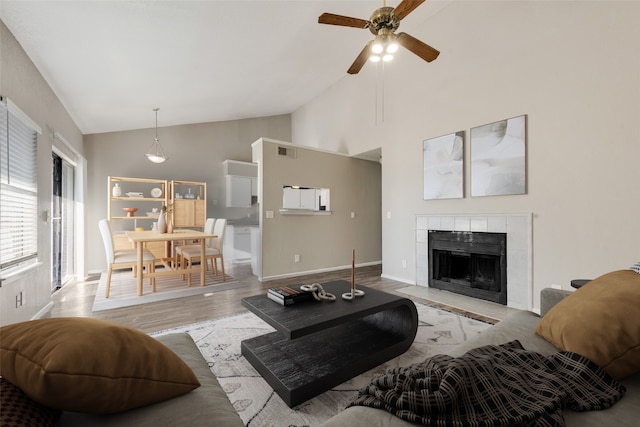 living room featuring ceiling fan, light wood-type flooring, high vaulted ceiling, and a tiled fireplace