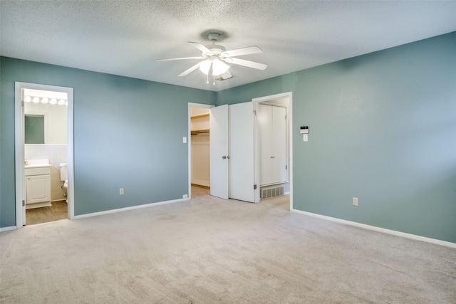 unfurnished bedroom featuring a walk in closet, ensuite bath, a textured ceiling, light carpet, and ceiling fan