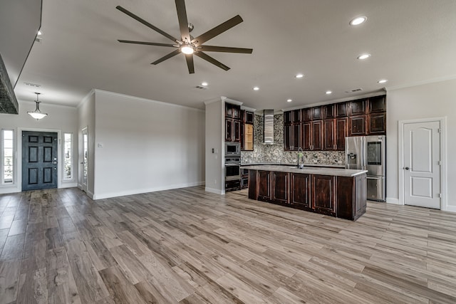 kitchen with an island with sink, wall chimney exhaust hood, stainless steel appliances, light hardwood / wood-style flooring, and decorative light fixtures