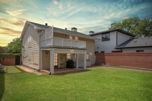 back house at dusk featuring a balcony, a lawn, and a patio