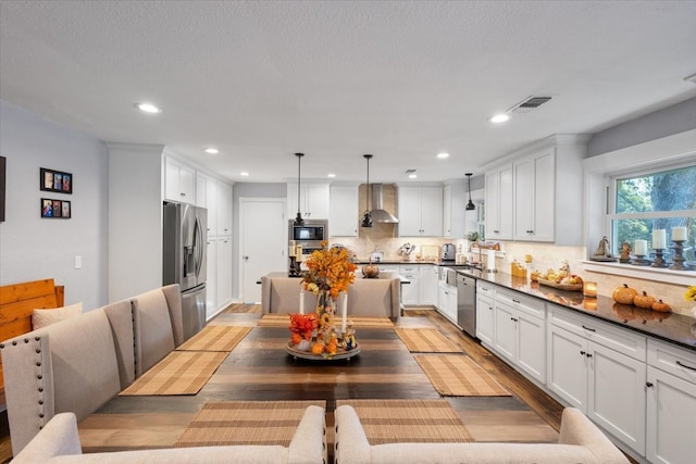 kitchen featuring pendant lighting, white cabinetry, wall chimney range hood, backsplash, and appliances with stainless steel finishes