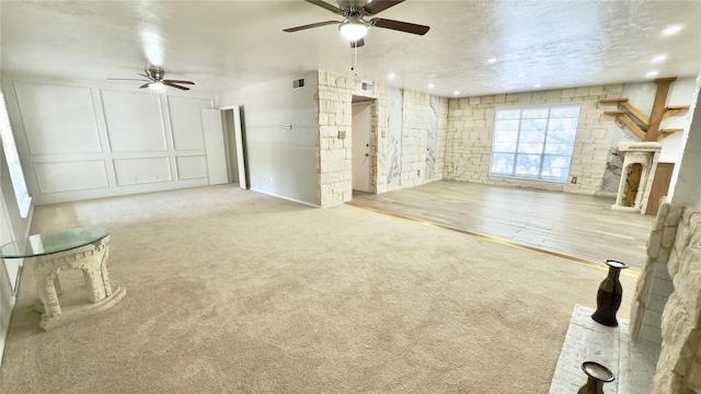 unfurnished living room featuring a textured ceiling, a stone fireplace, ceiling fan, and light colored carpet