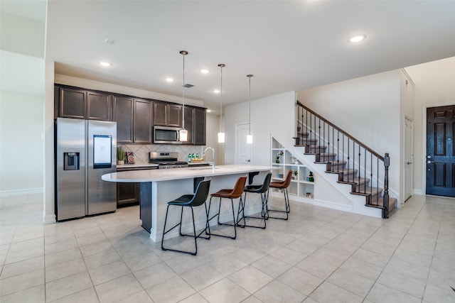 kitchen featuring appliances with stainless steel finishes, dark brown cabinets, light tile patterned floors, pendant lighting, and an island with sink