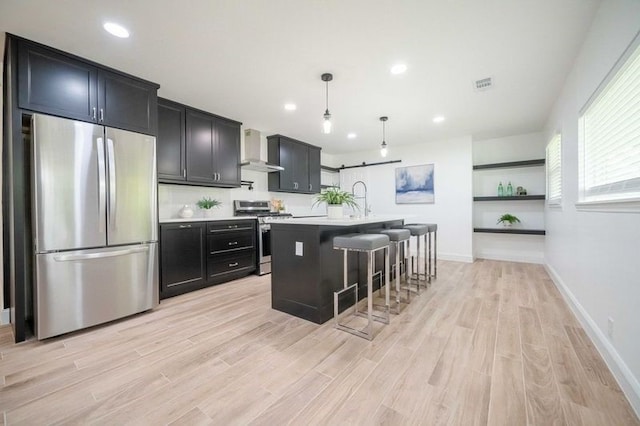 kitchen featuring appliances with stainless steel finishes, light hardwood / wood-style floors, a breakfast bar, wall chimney exhaust hood, and a center island with sink