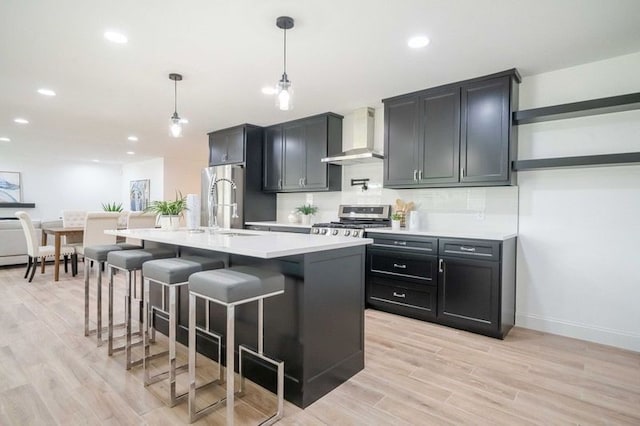 kitchen with an island with sink, pendant lighting, stainless steel appliances, light wood-type flooring, and wall chimney range hood