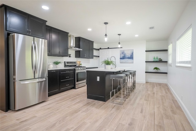 kitchen with a breakfast bar area, an island with sink, hanging light fixtures, wall chimney exhaust hood, and stainless steel appliances