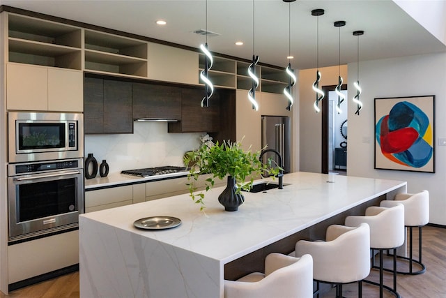 kitchen featuring appliances with stainless steel finishes, decorative light fixtures, decorative backsplash, a kitchen island with sink, and light stone counters