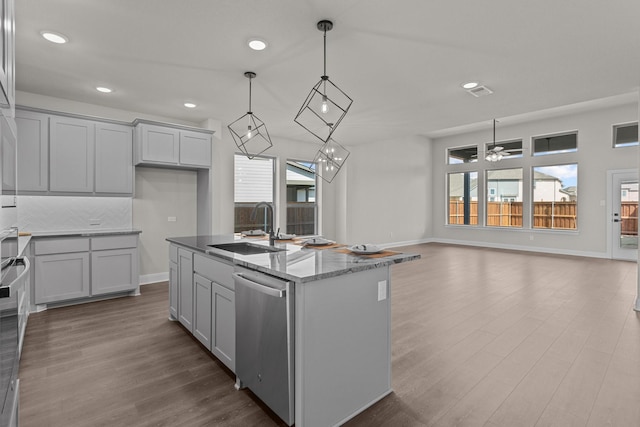 kitchen with light stone counters, decorative light fixtures, a center island with sink, stainless steel dishwasher, and a sink