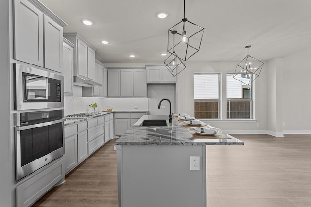 kitchen featuring an island with sink, appliances with stainless steel finishes, wood finished floors, gray cabinetry, and a sink