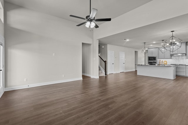 unfurnished living room with dark wood-style floors, ceiling fan with notable chandelier, baseboards, and stairs