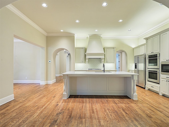 kitchen featuring appliances with stainless steel finishes, a kitchen island with sink, light hardwood / wood-style floors, and custom exhaust hood