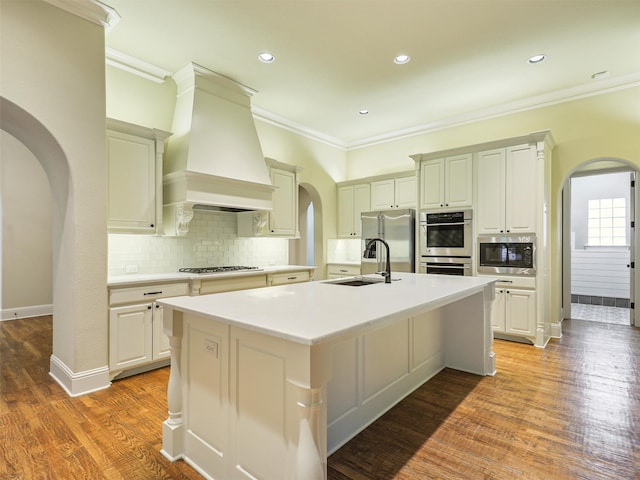 kitchen featuring custom exhaust hood, an island with sink, sink, stainless steel appliances, and hardwood / wood-style floors