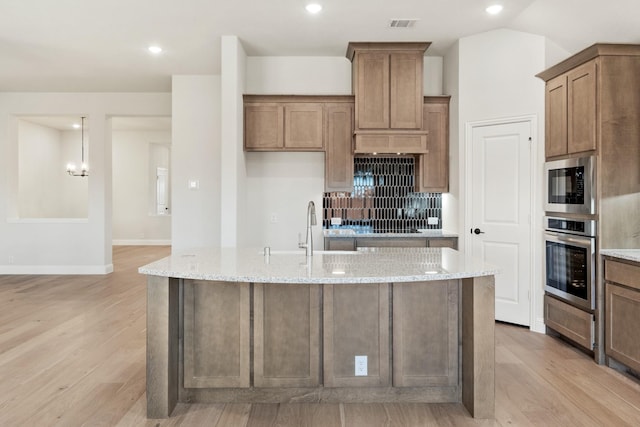 kitchen with a center island with sink, light stone counters, built in microwave, stainless steel oven, and a sink