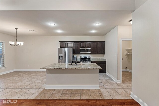 kitchen with light stone countertops, an inviting chandelier, a kitchen island with sink, dark brown cabinets, and appliances with stainless steel finishes