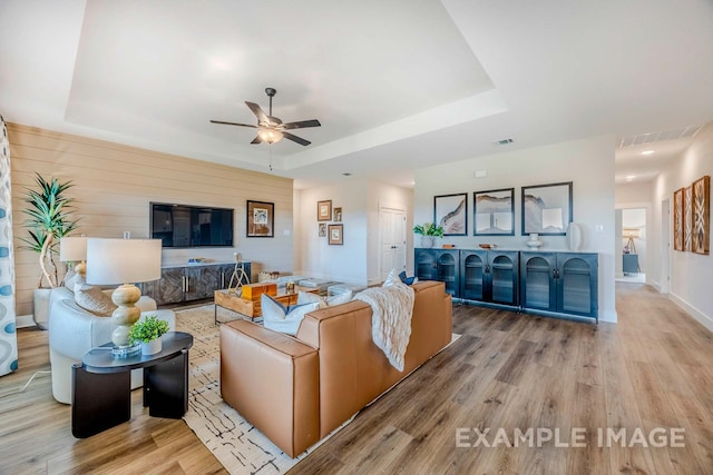 living room with ceiling fan, light hardwood / wood-style flooring, wooden walls, and a tray ceiling