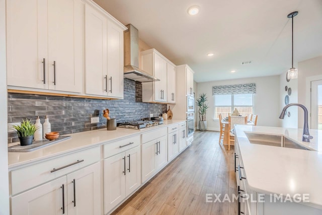 kitchen featuring white cabinetry, sink, wall chimney exhaust hood, hanging light fixtures, and light hardwood / wood-style flooring