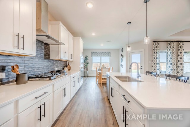 kitchen with appliances with stainless steel finishes, wall chimney exhaust hood, a kitchen island with sink, sink, and light hardwood / wood-style floors