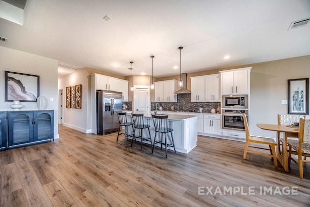 kitchen featuring white cabinetry, hanging light fixtures, stainless steel appliances, wall chimney range hood, and a center island with sink