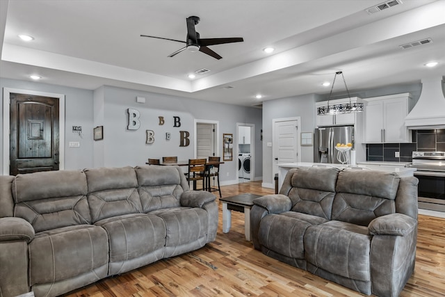 living room with light wood-type flooring, ceiling fan, a raised ceiling, and washer / clothes dryer