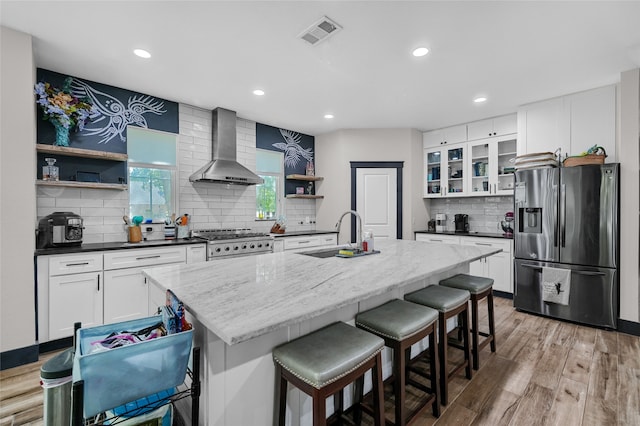 kitchen featuring wall chimney range hood, white cabinetry, a kitchen island with sink, light wood-type flooring, and stainless steel appliances