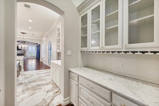 bar featuring crown molding, a fireplace, a notable chandelier, light stone counters, and white cabinetry