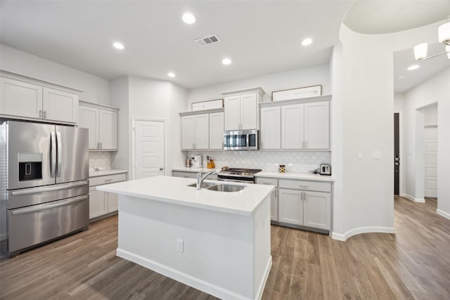 kitchen with hardwood / wood-style floors, sink, appliances with stainless steel finishes, a center island with sink, and backsplash