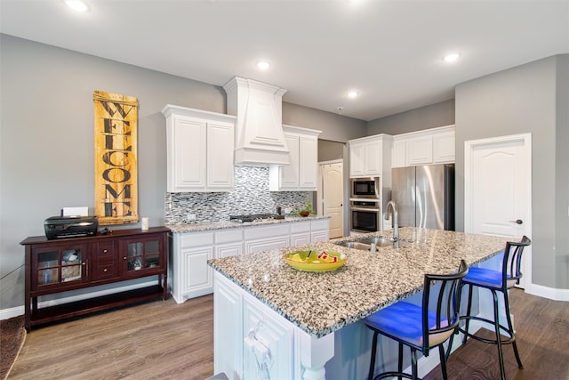 kitchen with sink, a center island with sink, white cabinetry, stainless steel appliances, and light stone countertops