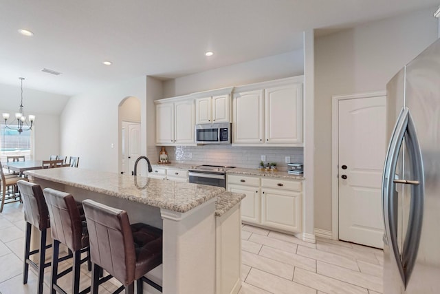 kitchen with white cabinetry, hanging light fixtures, stainless steel appliances, a center island with sink, and light stone countertops