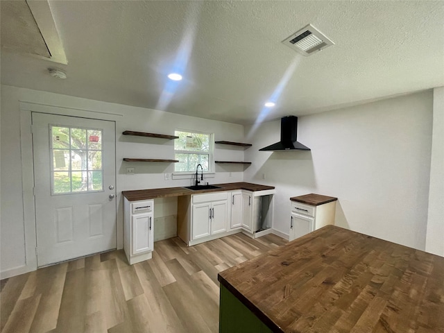 kitchen featuring plenty of natural light, sink, white cabinetry, wall chimney range hood, and butcher block countertops