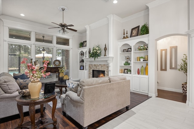 living room with a stone fireplace, ceiling fan, built in features, and ornamental molding