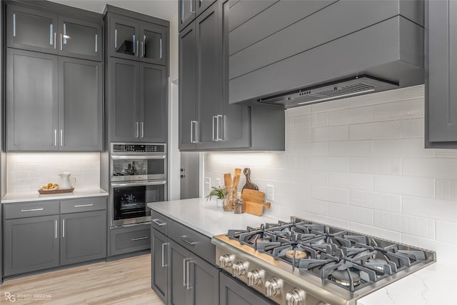 kitchen with appliances with stainless steel finishes, custom range hood, light hardwood / wood-style floors, light stone counters, and backsplash