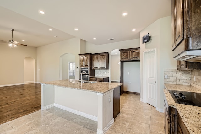 kitchen with light stone counters, ceiling fan, a center island with sink, black electric stovetop, and light wood-type flooring