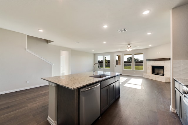 kitchen featuring dark hardwood / wood-style flooring, stainless steel dishwasher, sink, a stone fireplace, and an island with sink