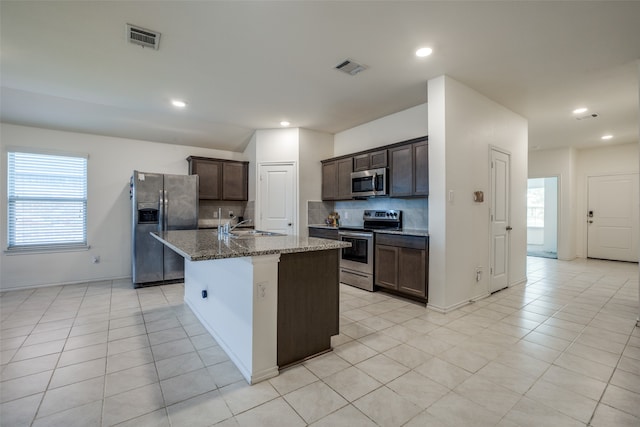 kitchen with light tile patterned flooring, dark brown cabinets, a kitchen island with sink, appliances with stainless steel finishes, and dark stone counters