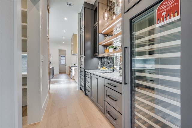 bar with light wood-type flooring, gray cabinetry, beverage cooler, light stone countertops, and decorative backsplash