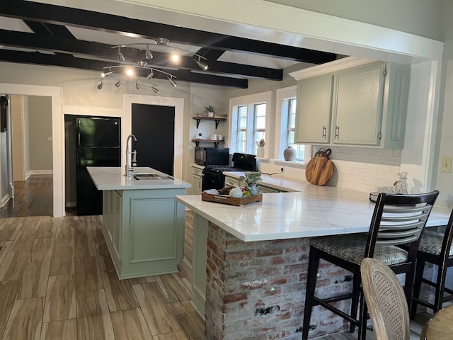 kitchen featuring black appliances, wood-type flooring, kitchen peninsula, a center island with sink, and beam ceiling