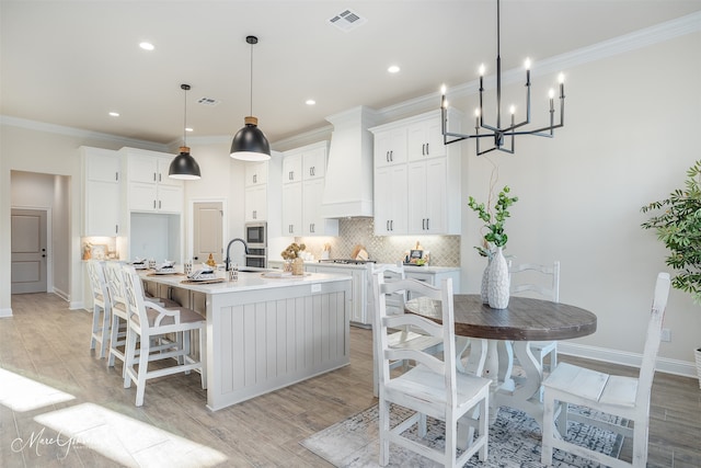 kitchen with pendant lighting, custom exhaust hood, a kitchen island with sink, light wood-type flooring, and white cabinetry