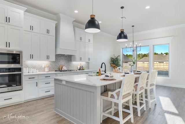 kitchen featuring white cabinetry, hanging light fixtures, stainless steel appliances, an island with sink, and custom exhaust hood