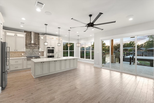 kitchen with wall chimney range hood, light hardwood / wood-style floors, pendant lighting, a center island with sink, and stainless steel appliances