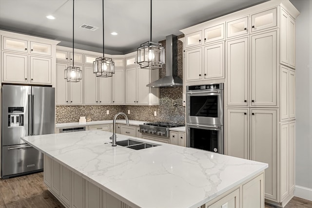 kitchen featuring hanging light fixtures, a kitchen island with sink, wall chimney range hood, appliances with stainless steel finishes, and light stone countertops