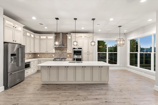 kitchen featuring an island with sink, stainless steel appliances, wall chimney range hood, and light hardwood / wood-style flooring