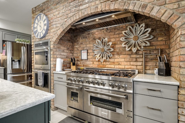 kitchen featuring range hood, appliances with stainless steel finishes, gray cabinets, and brick wall